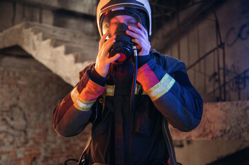 Ready, using a mask. Firefighter in professional equipment is indoors in abandoned building