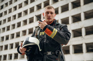 Conversation by walkie talkie. Professional firefighter is standing outdoors against big unfinished...