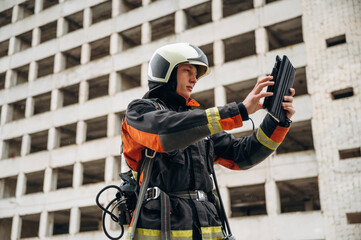Tablet in hands. Professional firefighter is standing outdoors against big unfinished building