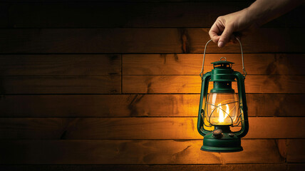 A dramatic photograph of a vintage kerosene lantern being held by a hand against a dark wooden background.