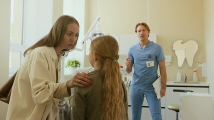 Medium shot of caring young mother talking to daughter in dental clinic encouraging child visiting dentist with friendly doctor in background - Powered by Adobe