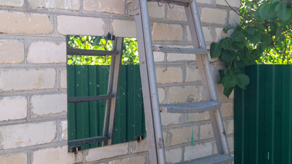 A metal ladder against a wall of white bricks and a square mirror. Mirror hung outside and metal ladder
