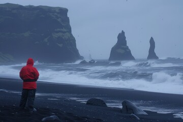 Person in a yellow raincoat stands on the black sand beach by the sea stacks on a misty day near Vik in Iceland