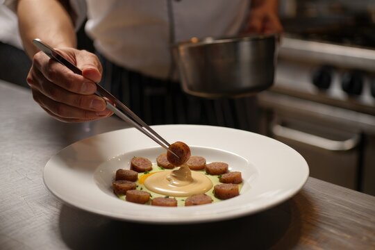 A chef carefully plates a gourmet sausage dish with tweezers and finesse