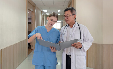 Two healthcare worker, a nurse in scrub and a doctor in a white coat, discuss patient file in a hospital corridor. They appear engaged and focused on the task at hand.