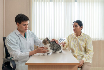 Young caucasian veterinarian in white gown examine a cat on the table while the owner look on, showcasing a caring and professional atmosphere in an animal clinic during daytime hour.