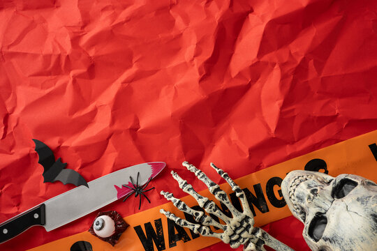 Halloween inspired composition with various decorations, including skeleton hand, warning tape, and knife, complemented by a festive red background