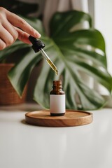 Hand holding dropper above amber bottle with blank label on wooden tray near monstera plant