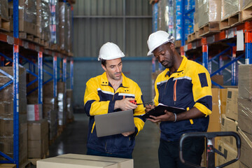 Two warehouse worker consult over a laptop and document clipboard while surrounded by stacked boxes and shelves, focusing on inventory management task in the afternoon.