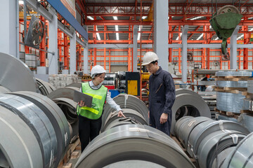 Two people are working in a factory. They are checking laptop computer within the metal sheet factory. Steel sheet roll stack are in the working area