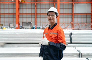 A worker in an orange jacket, hardhat, protective glove and safety glass stand confidently with finger thumbs up in front of a pile of material covered in plastic on the floor.