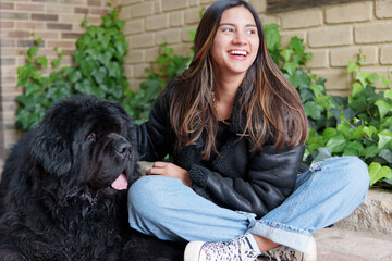 Happy young woman petting her large black newfoundland dog while sitting outdoors