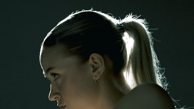 Side profile of a woman training on a stationary bike in a dark studio, with cinematic backlight wrapping her head and face in a glowing halo as she pedals with calm focus and inner strength