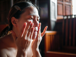 Beauty & Skincare, Asian woman applies facial cream sitting near a sunlit window She massaged her...