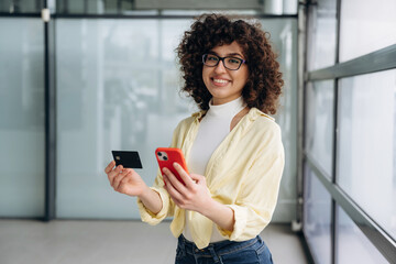 Credit card and phone in hands. Beautiful woman is standing indoors against blurred wall