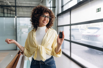Happy owner of a new car, holding keys. Beautiful woman is standing indoors against blurred wall
