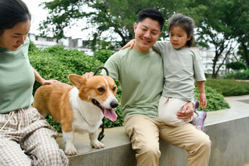 A father holds his daughter while petting his Corgi with his wife sitting beside him amidst a green...