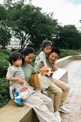 Smiling family with two kids and a pet dog sitting together on a concrete bench in a park, enjoying...