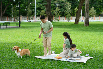 Father walks the family dog while mother and daughter sit on a picnic blanket in a peaceful green...