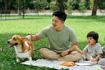 An Asian Father pets corgi while sitting with his young child on a picnic blanket in the greenery...