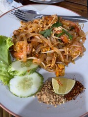 A plate of Pad Thai topped with shrimp, peanuts, and herbs, served with lime, chili flakes, cucumber slices, and lettuce, with a fork and spoon resting at the top of the plate.