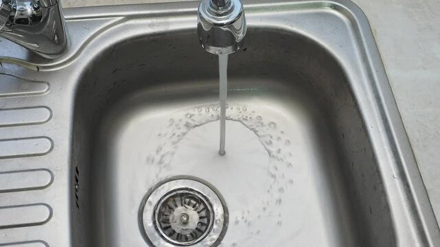 Clean water pours uselessly into a metal sink in the kitchen close-up waste of water
