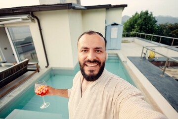 Happy man wearing bathrobe taking selfie by rooftop pool while holding cocktail