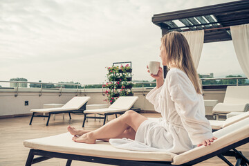 Woman in white bathrobe lying on sofa and relaxing with orange cocktail at the roof.