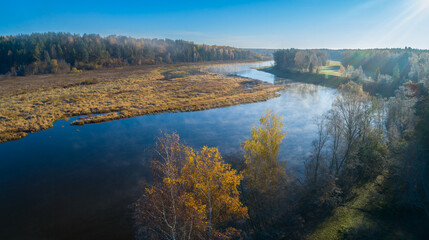 Morning sunrise view from above on river with trees on both banks and a misty haze in air. water is calm and trees are autumnal, sky is blue and clear.