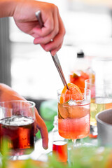 Close-up of a hand stirring a citrus cocktail with ice cubes in a glass, surrounded by ingredients...