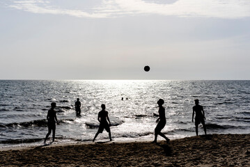 Silhouettes of young people playing with a ball on the beach at sunset with the sea shimmering in...