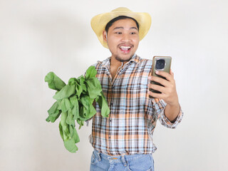 Modern Asian male farmer wearing a rattan hat holding a vegetable while looking at the smartphone...