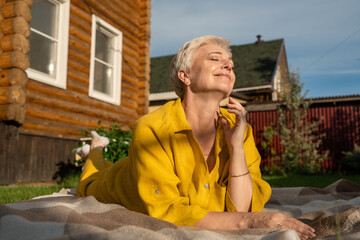 A serene scene of a mature woman dressed in yellow, leisurely relaxing on a blanket in her garden near house, enjoying sun