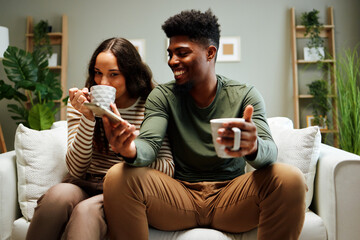 Young couple relaxing on sofa, drinking coffee and browsing smartphone