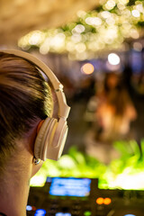 Rear view of DJ with white headphones at mixing console, blurred bokeh lights in background,...