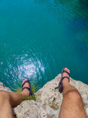 Man in sandals sitting on rocky cliff edge above clear blue sea, legs visible. Ideal for adventure,...