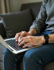Hands are typing on white laptop on dark gray sofa in living room, wearing black smartwatch. Technology, workspace, modern, comfort, innovation, casual, business