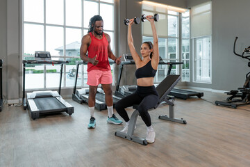 A personal trainer instruct a woman as she perform an overhead weight lift. They are in a well-lit...