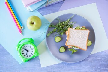 On the table is a school lunch, an alarm clock and stationery. A sandwich, an apple and a bottle of...