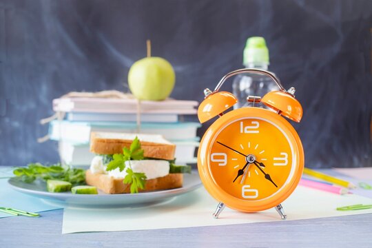 On the table is a school lunch, an alarm clock and stationery. A sandwich, an apple and a bottle of water. School lunch and textbooks.