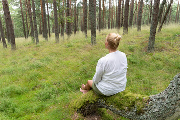 Peaceful blonde woman meditating in quiet pine forest. Natural green scenery, mossy tree, calm...