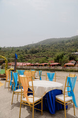 Outdoor event setup featuring decorated tables and chairs, set against a lush hillside backdrop. An...