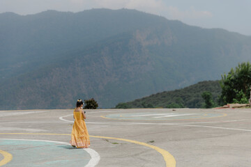Girl in yellow dress gazes at misty mountains from a helipad, enjoying scenic beauty and tranquil...