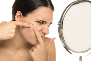 Woman squeezing pimple in front of mirror, painful expression, studio shot on white background.