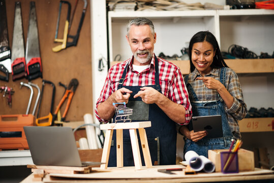 Senior Carpenter and Young Asian Partner Running Online Handcraft Wood Store, Using Laptop in Artisan Workshop for Digital Sales