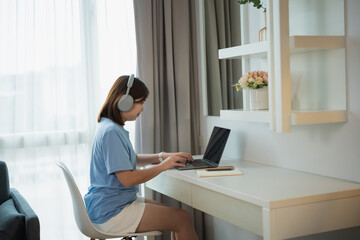 Young woman working on laptop at desk in bright room with headphones, enjoying remote work and study in a comfortable and modern environment