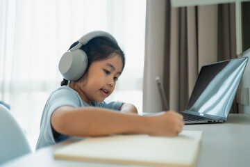 Young girl with headphones engaging in remote learning while writing notes at a desk with a laptop...