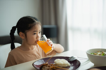 Young Girl Enjoying Bright Orange Drink with Healthy Meal on Table in Comfortable Indoor Setting, Natural Light Flowing Through Window