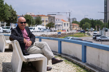 Elderly Spanish man in a blazer sitting on a bench in a small Portuguese town, looking at the beach