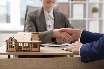 Real estate agent and client shaking hands at desk in office, focus on house model